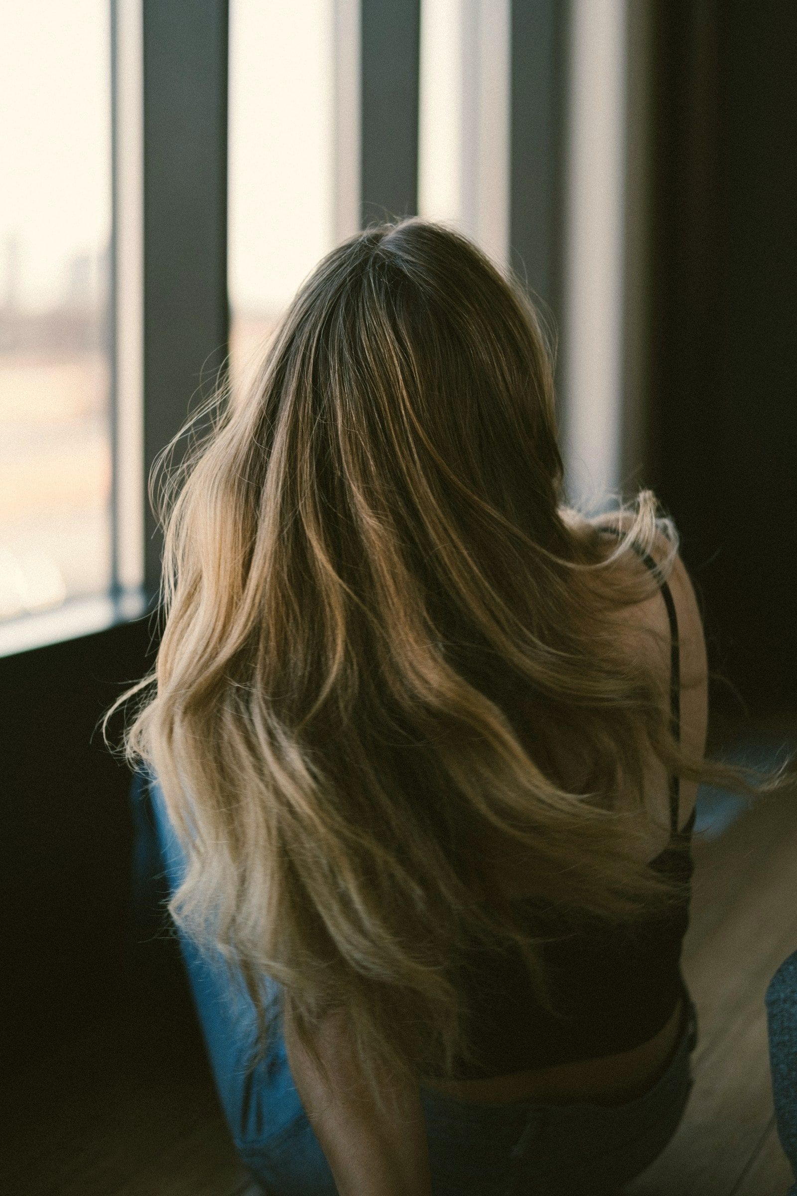 Langes Haar mit weichem, honigfarbenem Balayage, von hinten im Gegenlicht eines Fensters